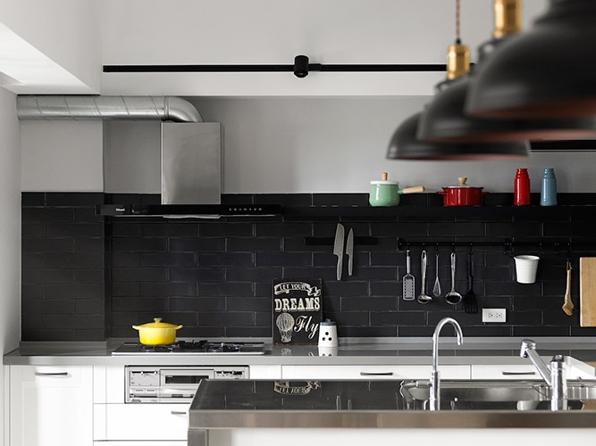 開放式的廚房安排符合屋主平時少油煙的烹調習慣。黑色的仿磚材質與燈飾、軌道燈相互呼應,搭配刻意外露的排油煙管將工業風元素輕鬆帶入,為簡約的生活畫面增添些許亮點。