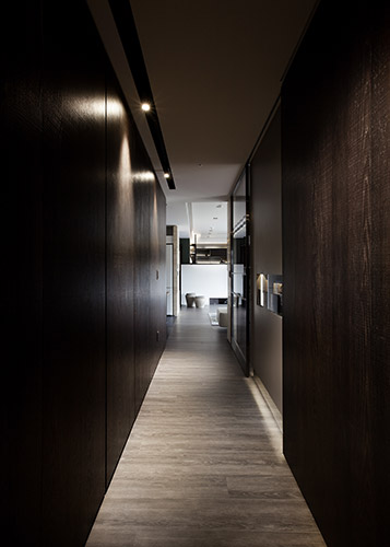 在公共空間過渡至私領域的廊道以深色刀痕鋼刷木皮的深色基調圍塑成熟穩重的氛圍，點綴以大地色系，做為公私領域的歇心轉折。